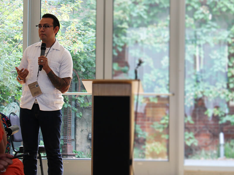 Photo of Juan Ramirez standing to the side of the podium and gesturing while speaking into a mic