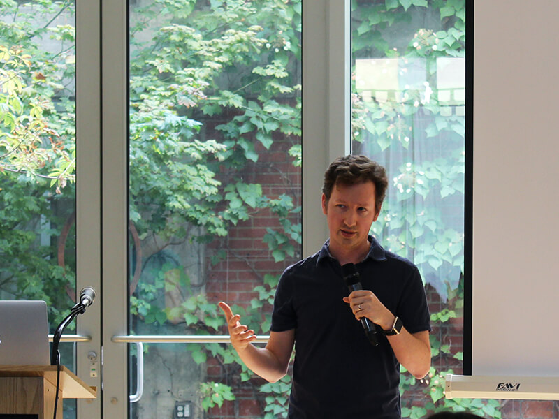 Photo of Nick standing and gesturing in front of a projector screen while holding a mic